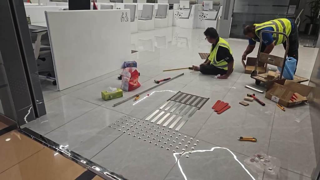 Two workers in safety vests assembling materials on a tiled office floor, with tools and items spread out around them.