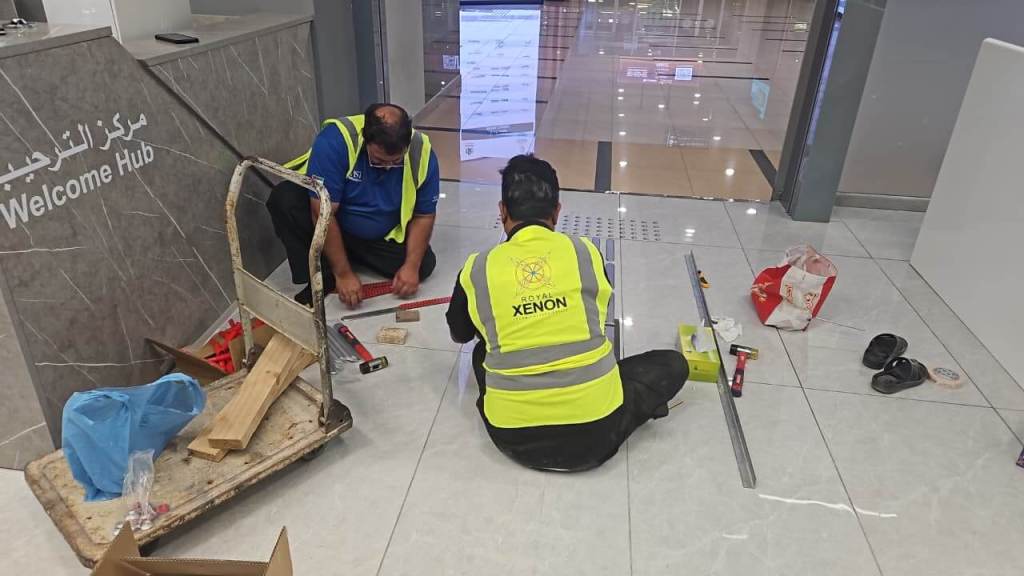 Two workers in safety vests are sitting on the floor of a modern space, engaged in a construction task near a reception area labeled 'Welcome Hub'. They are using tools and materials spread out on the floor, with a cart and various supplies nearby.