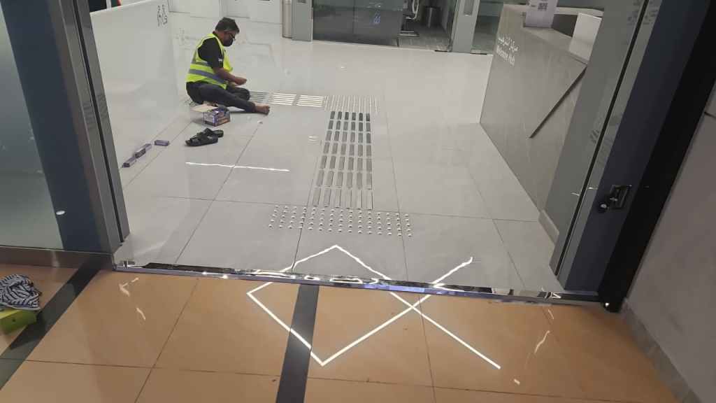 A worker in a yellow safety vest kneels on the floor, installing floor tiles in a modern indoor space with reflective surfaces and bright lighting.
