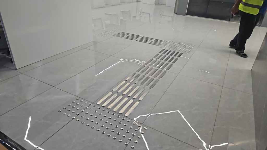 A polished marble floor featuring an arrangement of metallic strip and dot markers for tactile guidance, with a person in a safety vest standing nearby.