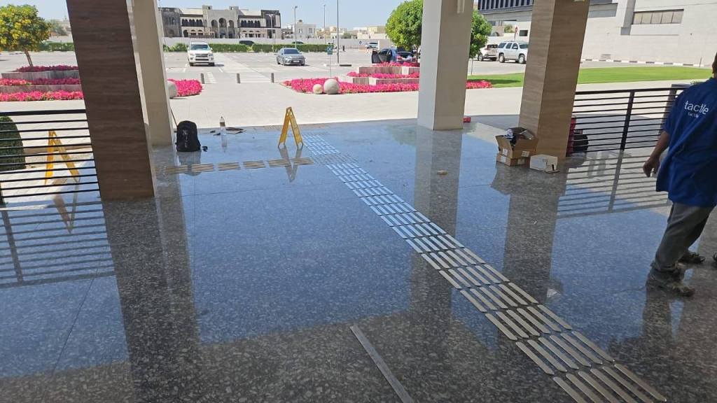A view of a polished stone floor in a building entrance, with wet areas and caution signs visible. A worker in a blue shirt is walking, and boxes are scattered nearby. Brightly colored flowers are planted outside the entrance.