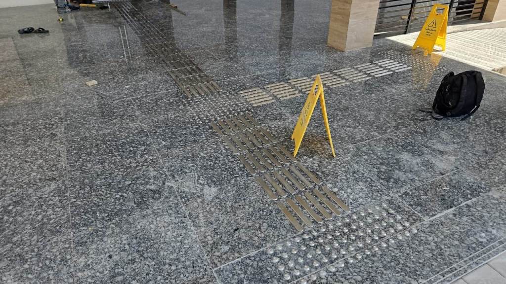 A tiled floor with a textured pattern for tactile guidance, featuring caution signs and a black backpack nearby.
