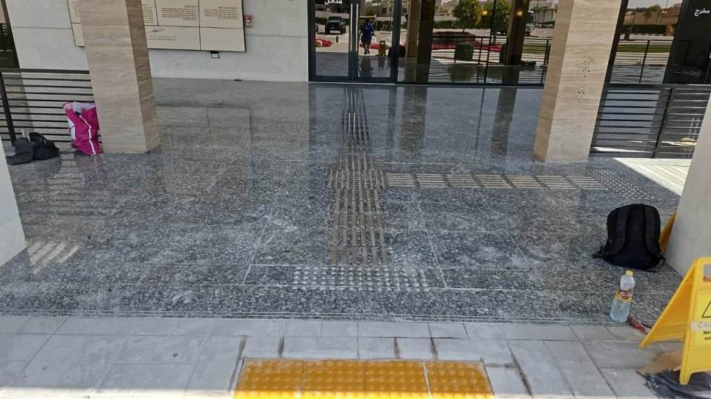 Interior view of a polished granite floor with tactile paving for accessibility. A pink bag and a black backpack are placed on the floor, and a caution sign is visible on the right.