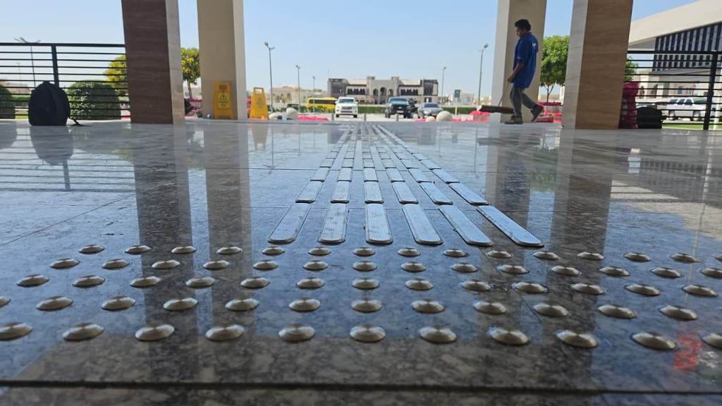A close-up view of a textured floor with tactile paving strips and raised dots for visually impaired individuals, taken from a low angle, with a person walking in the background.