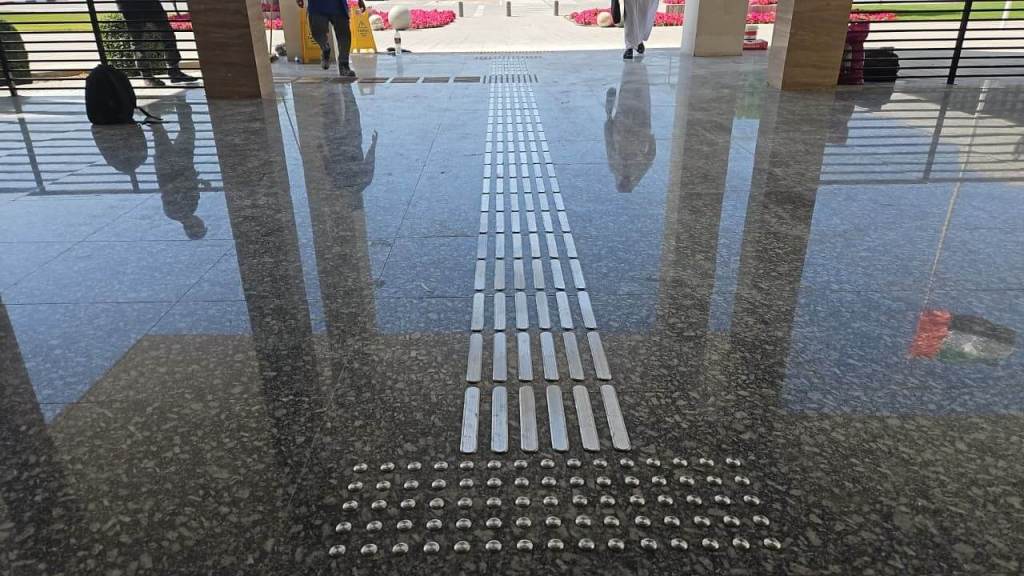 A polished stone floor featuring a tactile guide strip with alternating smooth and textured elements, leading towards an outdoor area with greenery and flowers in the background.