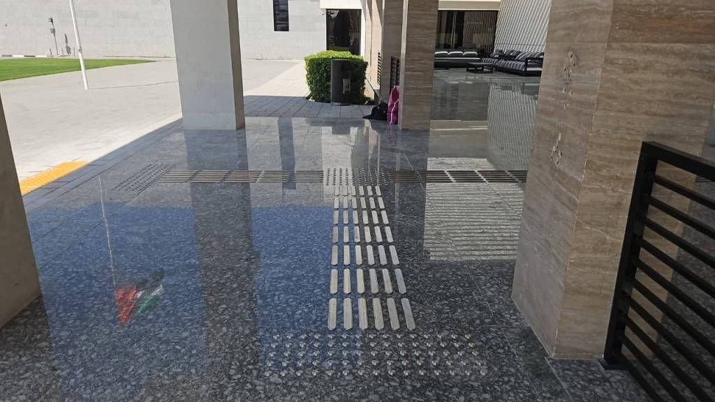A polished granite floor with tactile pathway markers and a reflective surface, leading towards a grassy area and buildings in the background.