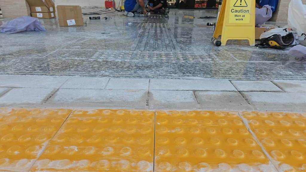 A construction site featuring textured yellow tactile paving on the ground, with workers in the background and a caution sign indicating 'Men at work'.