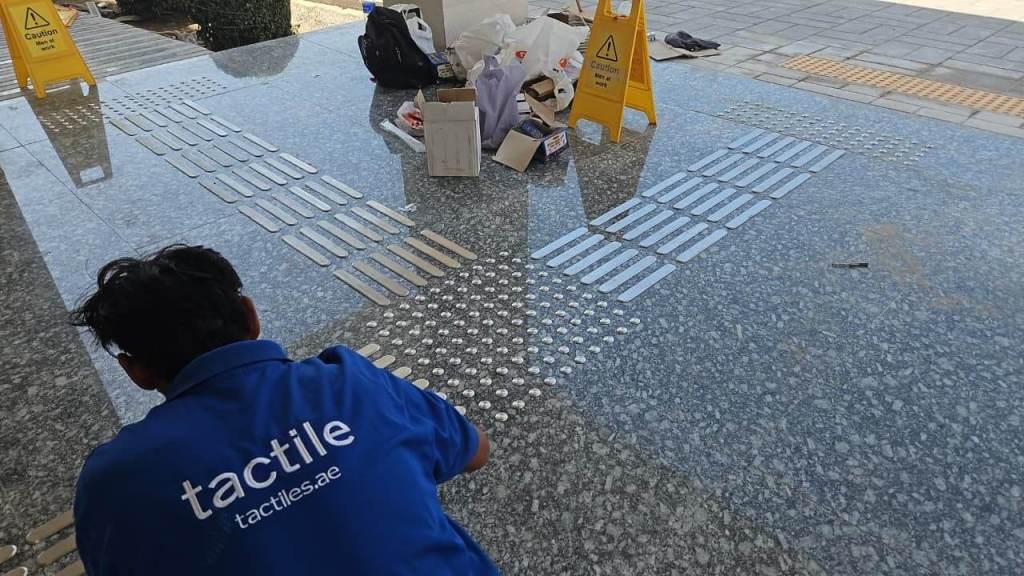 A person in a blue shirt is working on installing tactile tiles on a smooth granite surface. Caution signs are visible on the ground, and various materials are scattered nearby.