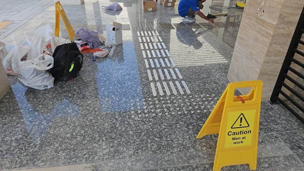 A construction area with a caution sign that reads 'Caution Men at work,' scattered bags and tools, and a worker installing flooring materials.