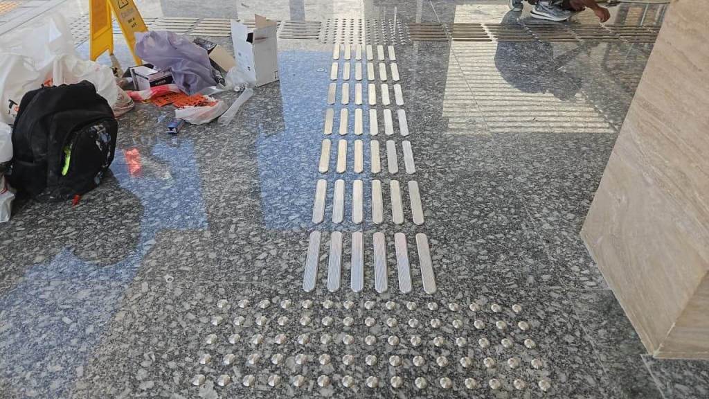 A close-up view of a tiled floor with shiny metallic tactile strips and raised dots, surrounded by scattered items including a backpack and some discarded materials.