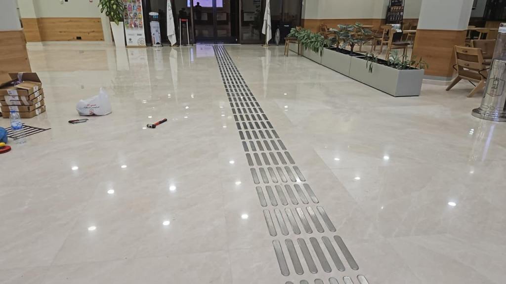 Interior of a spacious hallway with shiny marble flooring and a linear drainage system. There are cardboard boxes and tools scattered on the floor, and a plant box along one side.