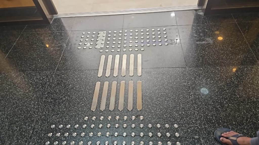 Close-up view of a textured floor featuring metal tactile indicators and strips, designed for guidance and accessibility near an entrance.