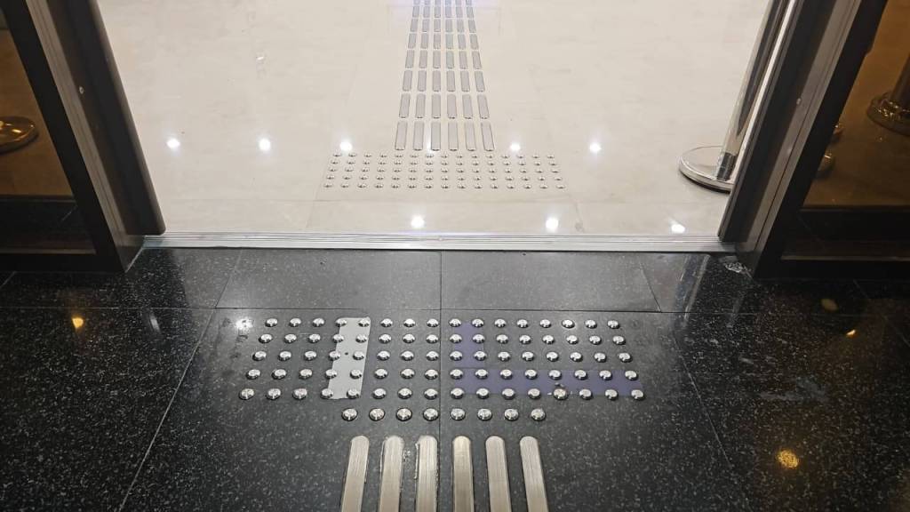 Close-up view of textured flooring at an entrance, featuring tactile indicators for visually impaired individuals. The surface transitions from black stone to a lighter tile, with metal strips and dots arranged on the ground.