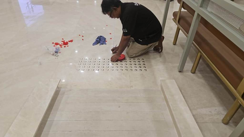 A man kneeling on a polished floor, placing small round objects in a patterned layout. Surrounding him are a few scattered red items and a blue cloth, with a staircase visible in the foreground.