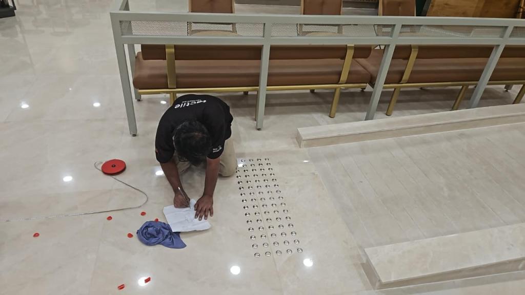 A person measuring and marking lines on a marble floor in an indoor setting, with benches in the background and tools scattered nearby.