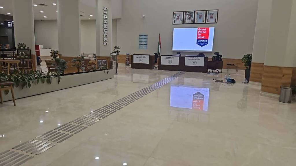 Interior view of a modern building featuring a Starbucks coffee shop, comfortable seating, and a large screen displaying a certification for 'Great Place to Work'. The floor is polished and has a drainage line running through it.