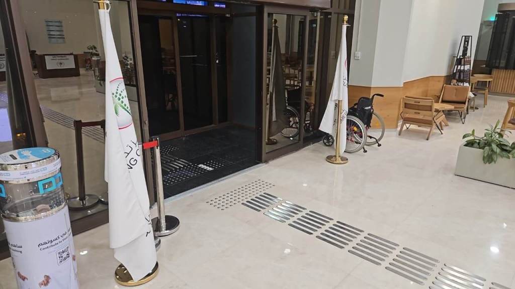An entryway featuring glass doors, accessible signage, and a wheelchair near the entrance. Two flags are positioned beside the doors, and there are chairs and a plant in the lobby area.