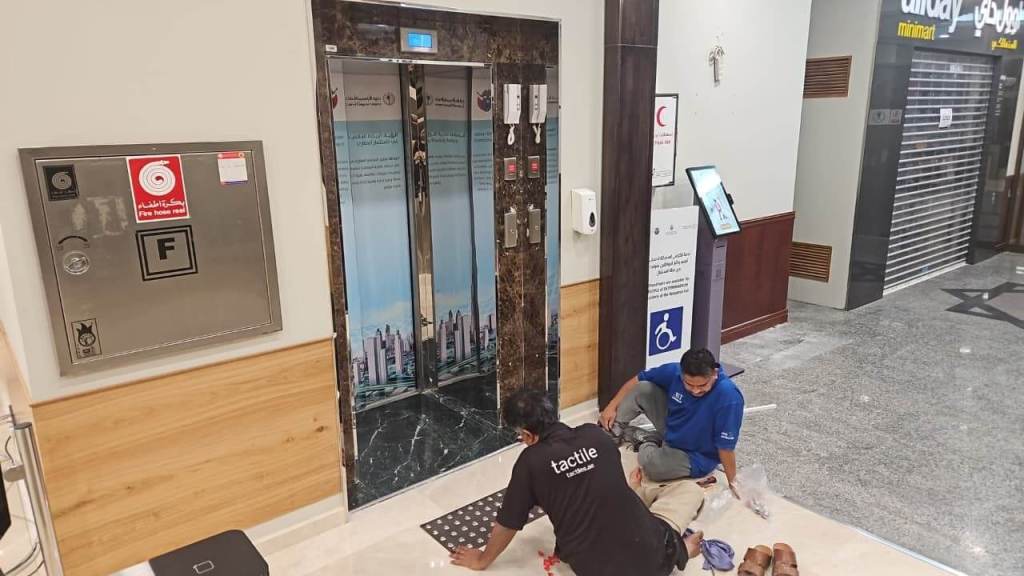 Two workers performing maintenance on the floor near an elevator in a modern building lobby, with signage and accessibility features visible.