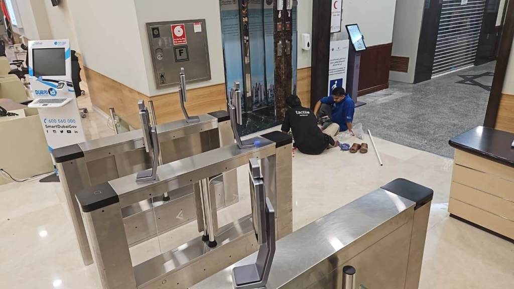 Inside a modern building, two individuals work on the floor near turnstiles, with a digital kiosk and closed door visible in the background.