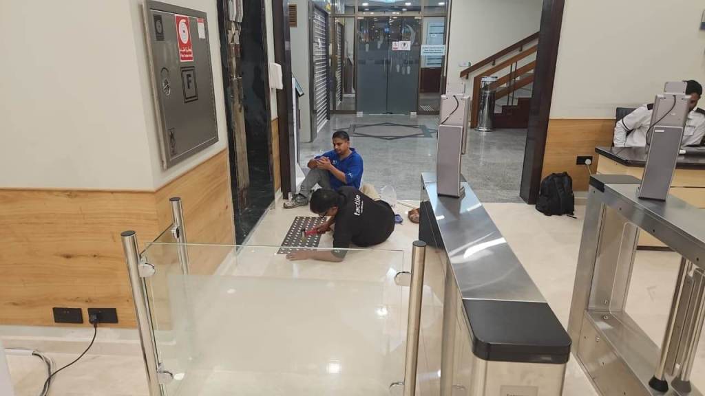 Two technicians working on a floor installation in a modern lobby area with turnstiles and a staircase in the background.
