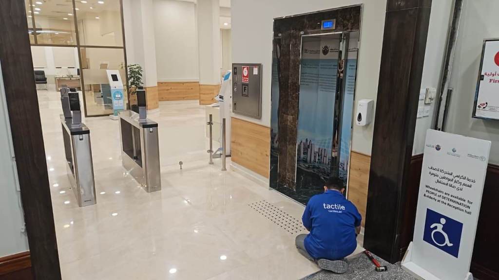 A maintenance worker in a blue shirt is working on the ground near an elevator in a modern building lobby. The area features automated turnstiles, an elevator, a hand sanitizer station, and a sign indicating accessibility services.