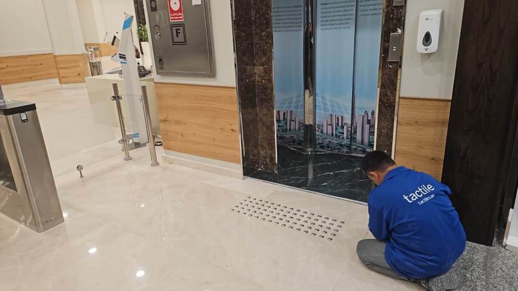A worker in a blue 'tactile' shirt kneels on the floor next to an elevator entrance, inspecting the area. The elevator doors feature an image of a city skyline, and there is a hand sanitizer dispenser mounted on the wall.