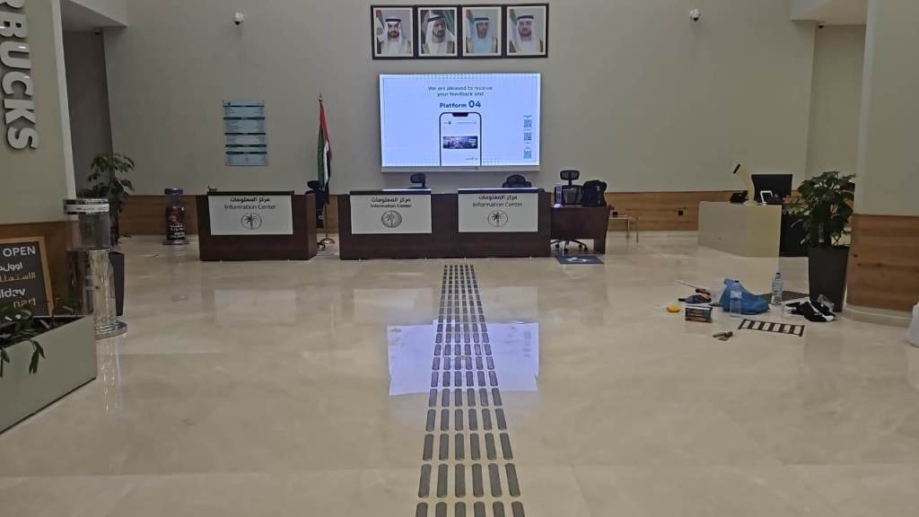 Interior of an information center featuring a large screen displaying information, with chairs and tables arranged in front. The space includes elements like plants and cleaning supplies scattered on the floor.