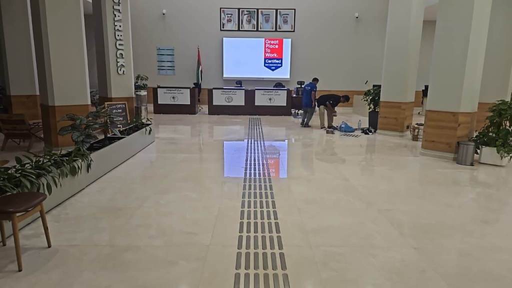 Interior view of a reception area with a Starbucks sign, featuring a polished floor, decorative plants, and workers preparing the space. A screen displays a 'Great Place to Work' certification.