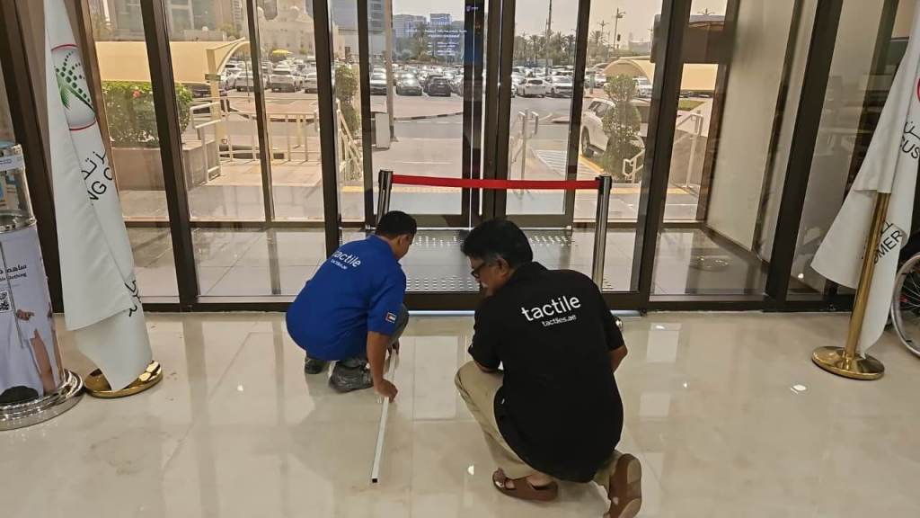 Two individuals working together on a floor installation project inside a building entrance, one measuring with a tool while the other observes, with a red barrier in the background.