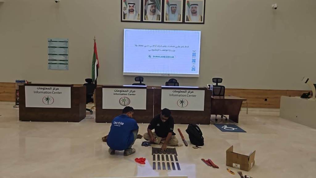 A man working on the floor of an Information Center while another person is assembling materials. In the background, there are flags and a display screen showing information.