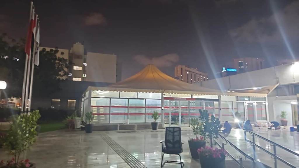 Night view of a building with a tent-like structure, surrounded by potted plants and flags. The area is illuminated, showing reflections on the floor.