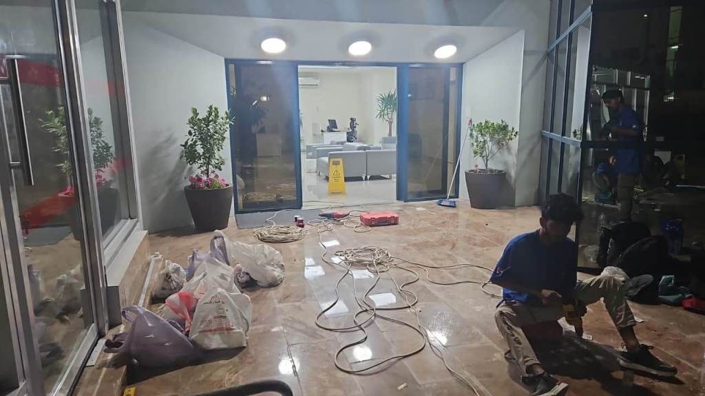 An interior space under renovation with a worker sitting on the floor using a tool. There are construction materials and bags scattered on the floor, along with potted plants and a wet floor sign in the background.