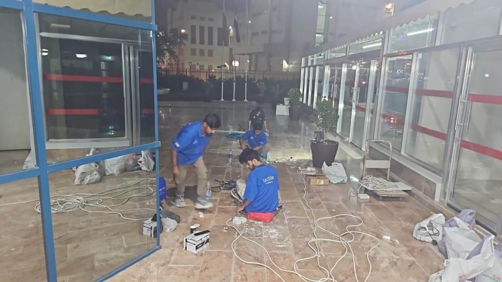 Workers in blue shirts assembling equipment at night in a commercial space, with tools and materials scattered on the tiled floor.
