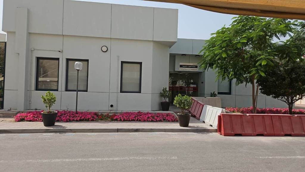 Building exterior with large windows, surrounded by flower beds and greenery, featuring flower pots and construction barriers in front.