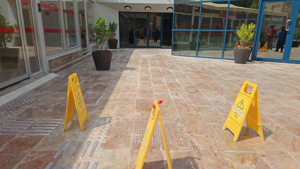 A tiled outdoor entryway with yellow caution signs indicating wet floors, decorative plants in pots, and glass doors in the background.