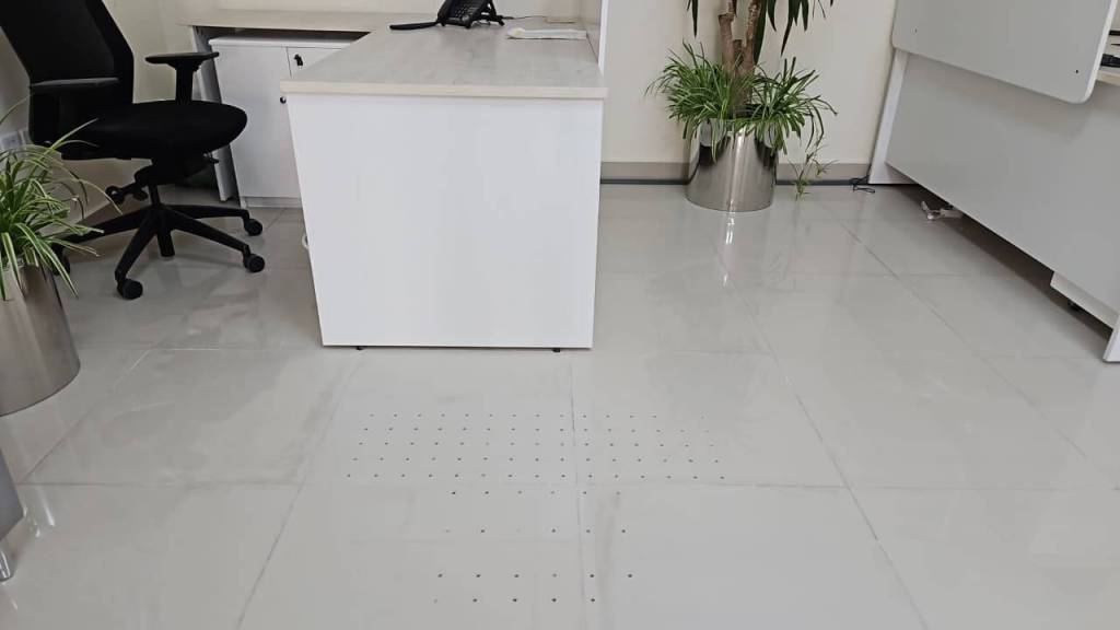 A modern office space featuring a white desk, black chair, and potted plants, with shiny tiled flooring that has a dotted pattern.
