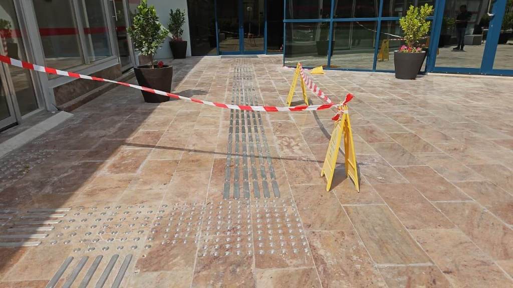 A partially blocked outdoor area with red and white caution tape and yellow warning signs. The flooring features tactile guide strips, and potted plants are visible in the background.