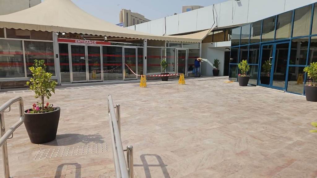 Outdoor entrance to a building with sliding glass doors, a large canopy above, and safety cones placed on the ground. Potted plants are positioned near the entrance.