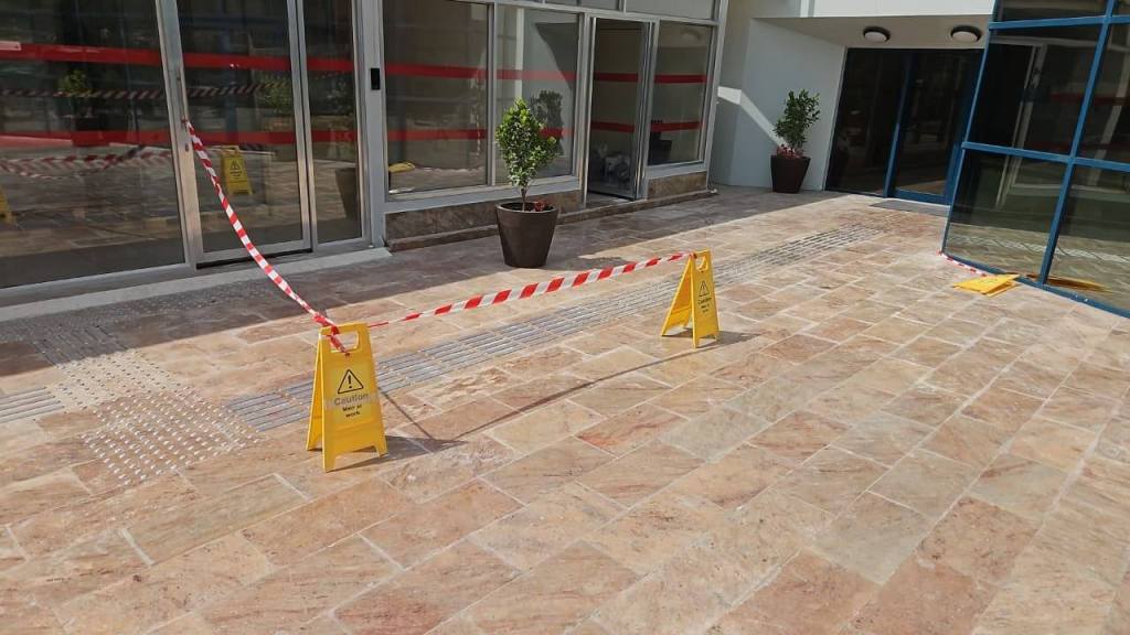 Floor area with caution signs indicating a wet surface, marked by red and white barrier tape, near a glass entrance.