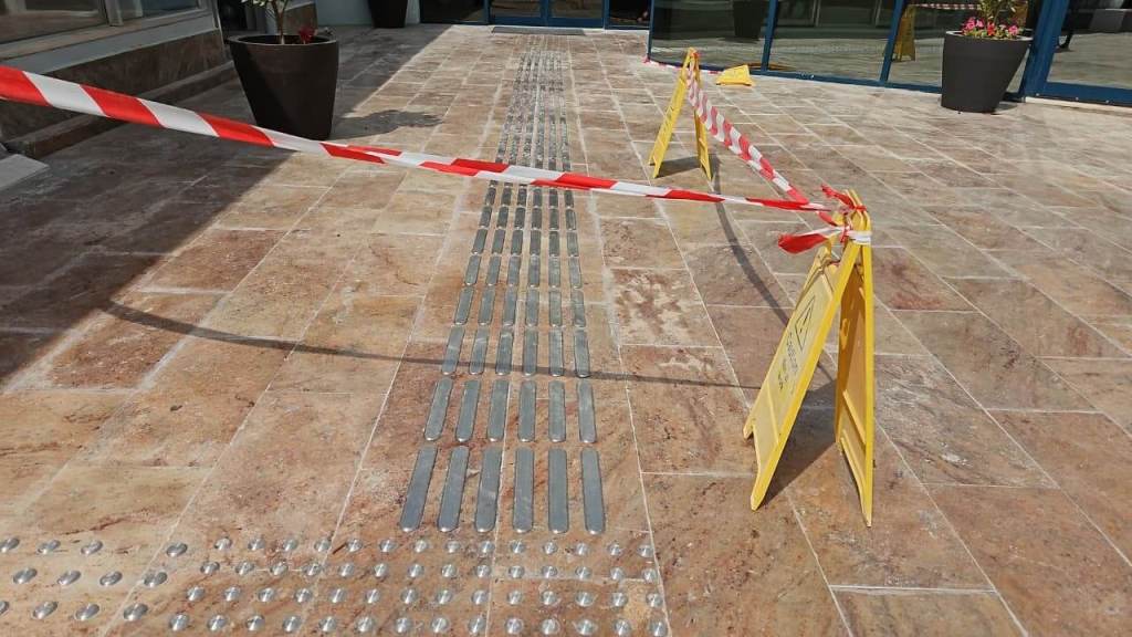 A view of a tiled walkway with caution tape and warning signs indicating a restricted area, featuring tactile paving for accessibility.