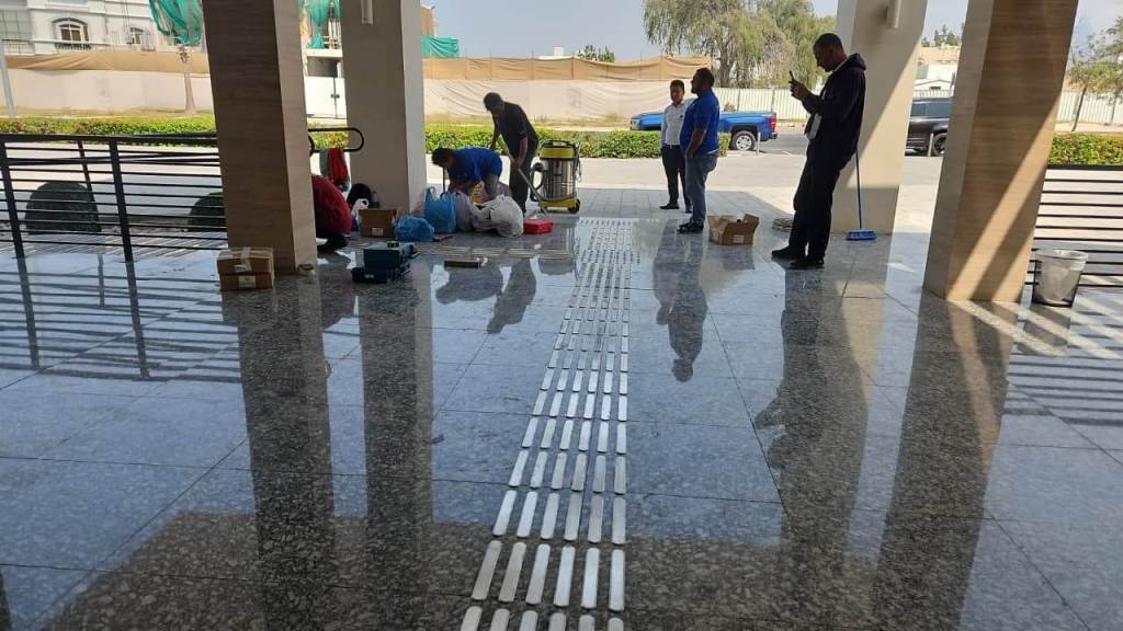 A group of people working in a modern entrance lobby with shiny marble floors. Some individuals are engaged in tasks on the floor, while others stand nearby, observing or using their phones.