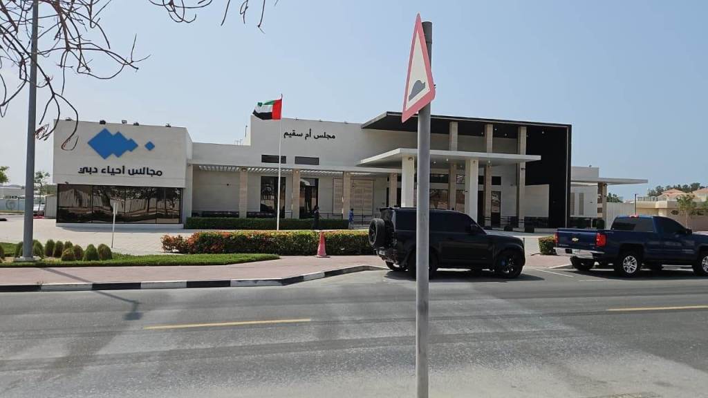 A modern building featuring a large entrance with a flat roof, labeled in Arabic and displaying the UAE flag. Several vehicles parked in front and a street sign visible.