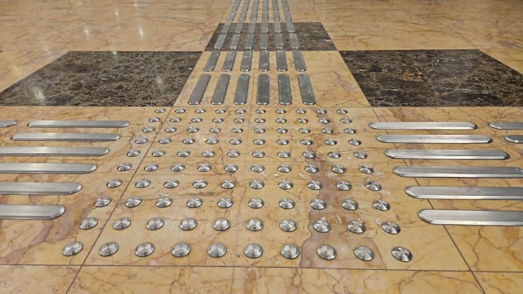 A close-up view of a textured floor with tactile warning strips and domed indicators, featuring a mix of metallic and smooth surfaces.