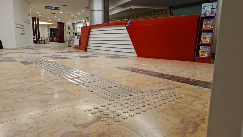 Interior of a modern reception area with a red front desk, featuring tactile guidance indicators on the floor and seating in the background.