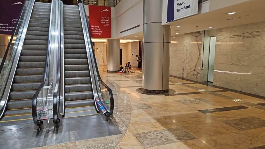 A spacious interior view featuring two escalators, polished marble flooring, a large column, and colorful signage on the walls. A few people are seated on the floor nearby.