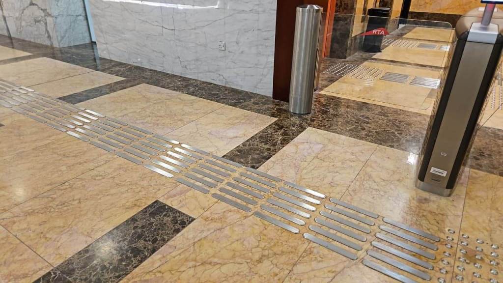 An interior floor view showing a decorative marble surface with metallic tactile paving strips for accessibility, located near automated turnstiles in a building entrance.