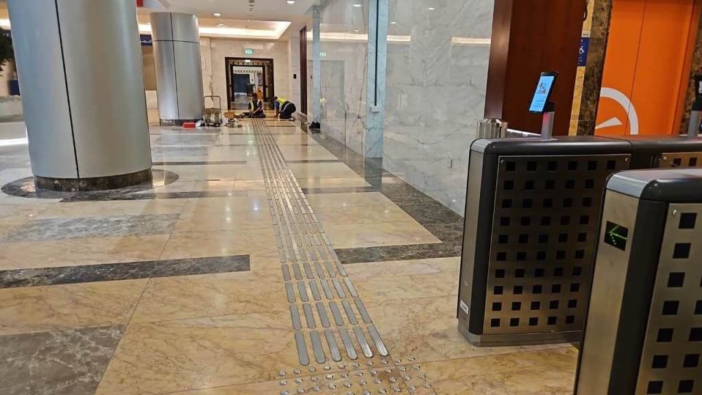 Interior of a modern building with polished marble flooring, featuring a tactile guide path on the floor, security turnstiles on the right, and two workers in yellow vests performing maintenance in the background.