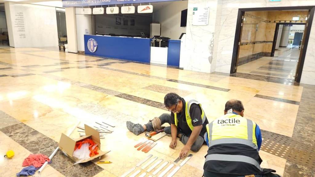 Two workers wearing safety vests are sitting on the floor of a lobby, installing tactile strips. A box with tools and materials is nearby.