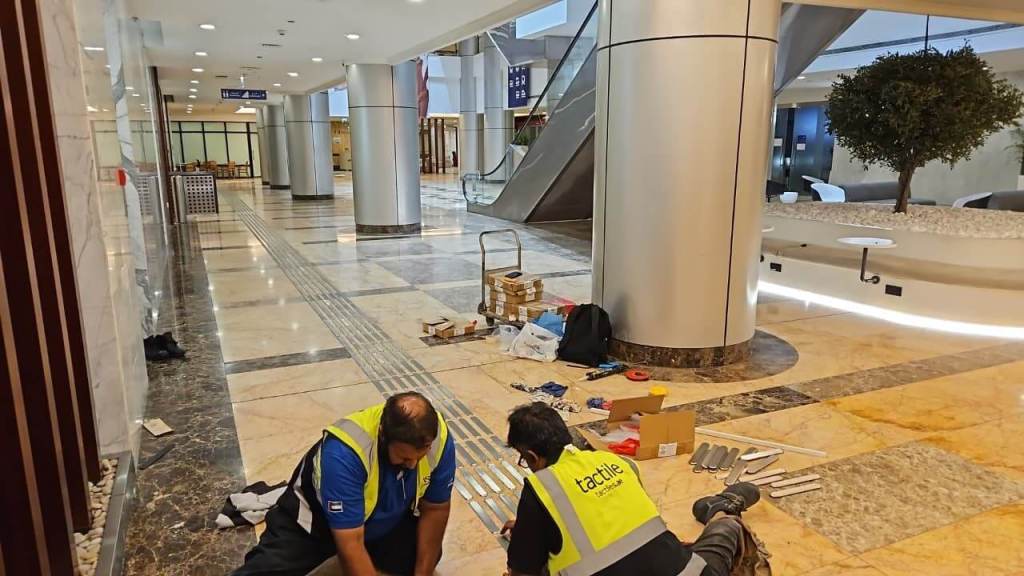 Two workers in reflective vests are seated on the floor of a modern lobby, assembling equipment. Tools and materials are scattered around them, and an escalator and decorative plants are visible in the background.