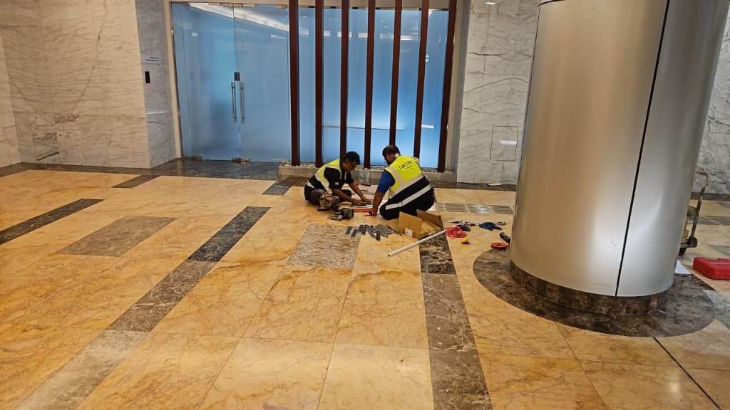 Two workers in safety vests assembling equipment on marble flooring in a lobby, with tools scattered around and glass doors in the background.